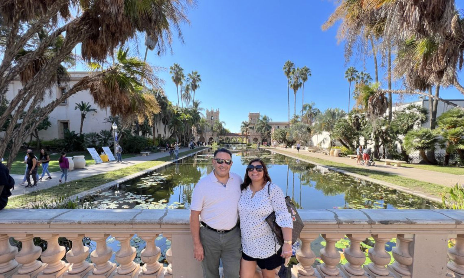Two people smiling at a stone balustrade overlooking a long reflecting pond with lily pads, tall palm trees and Spanish‑style architecture under a bright blue sky — sunny outdoor courtyard scene.