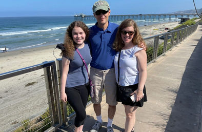 Three smiling people pose on a sunny coastal boardwalk overlooking a sandy beach and ocean pier with gentle waves under a clear blue sky.