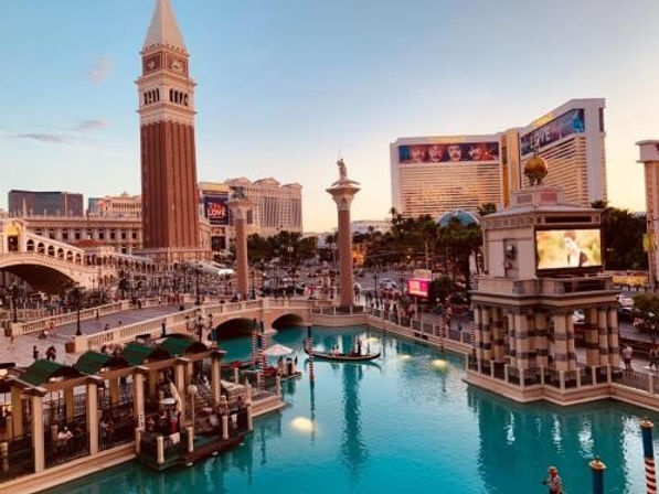 Las Vegas Strip evening scene with turquoise resort canal, gondola riders, Venetian-style bell tower and arched pedestrian bridges against a glowing skyline.