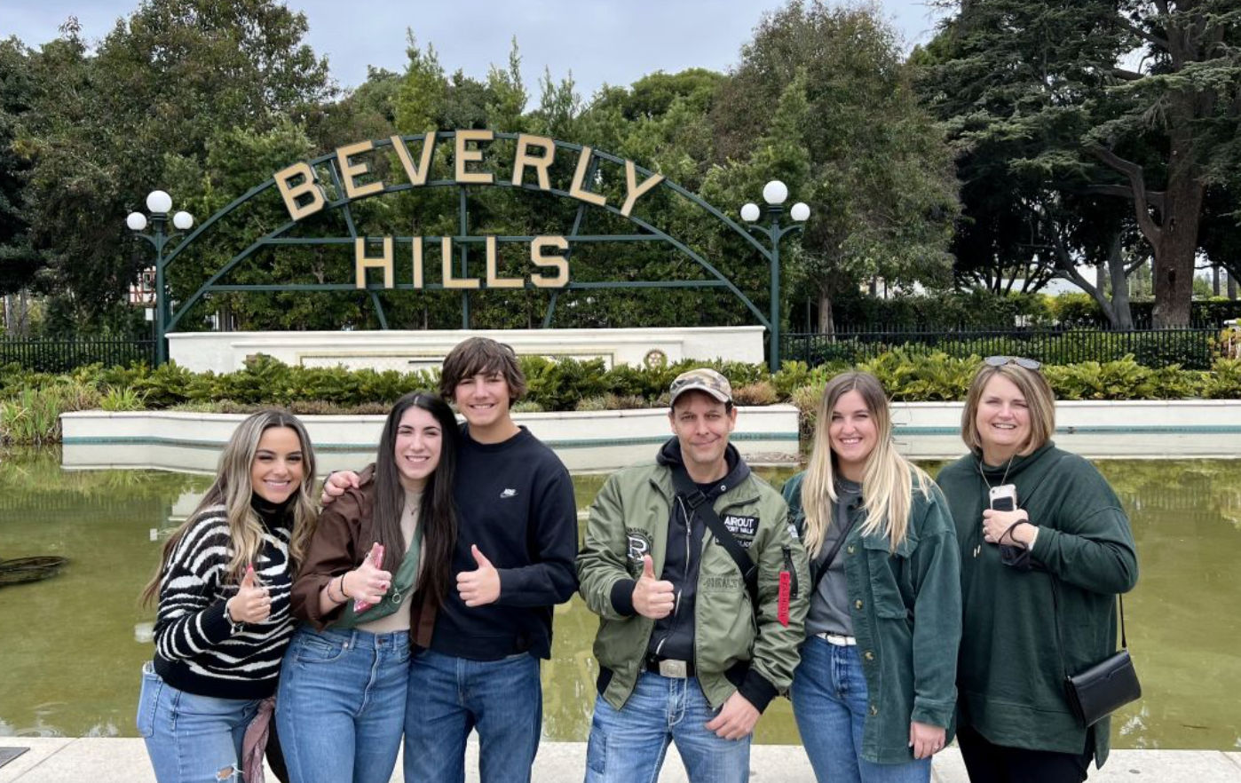 Six people smiling and giving thumbs-up in front of the Beverly Hills sign above a reflecting pool, with park trees and an overcast sky in the background.