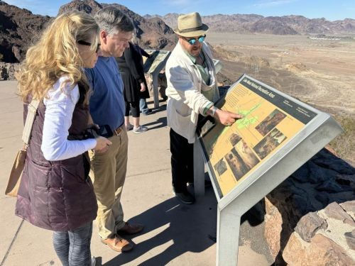 Three visitors at a sunny desert overlook reading an interpretive map; a man in a hat points to the sign with rocky mountains and a wide arid valley beyond.