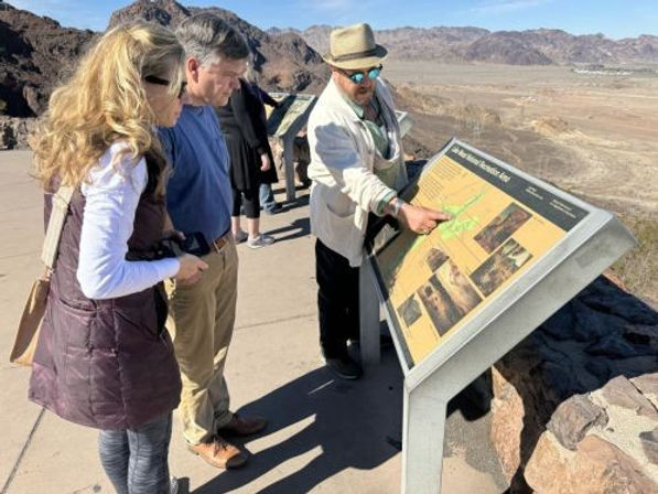 Three visitors at a sunny desert overlook reading an interpretive map; a man in a hat points to the sign with rocky mountains and a wide arid valley beyond.