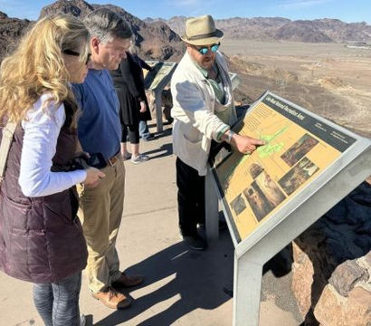 Three visitors at a sunny desert overlook reading an interpretive map; a man in a hat points to the sign with rocky mountains and a wide arid valley beyond.