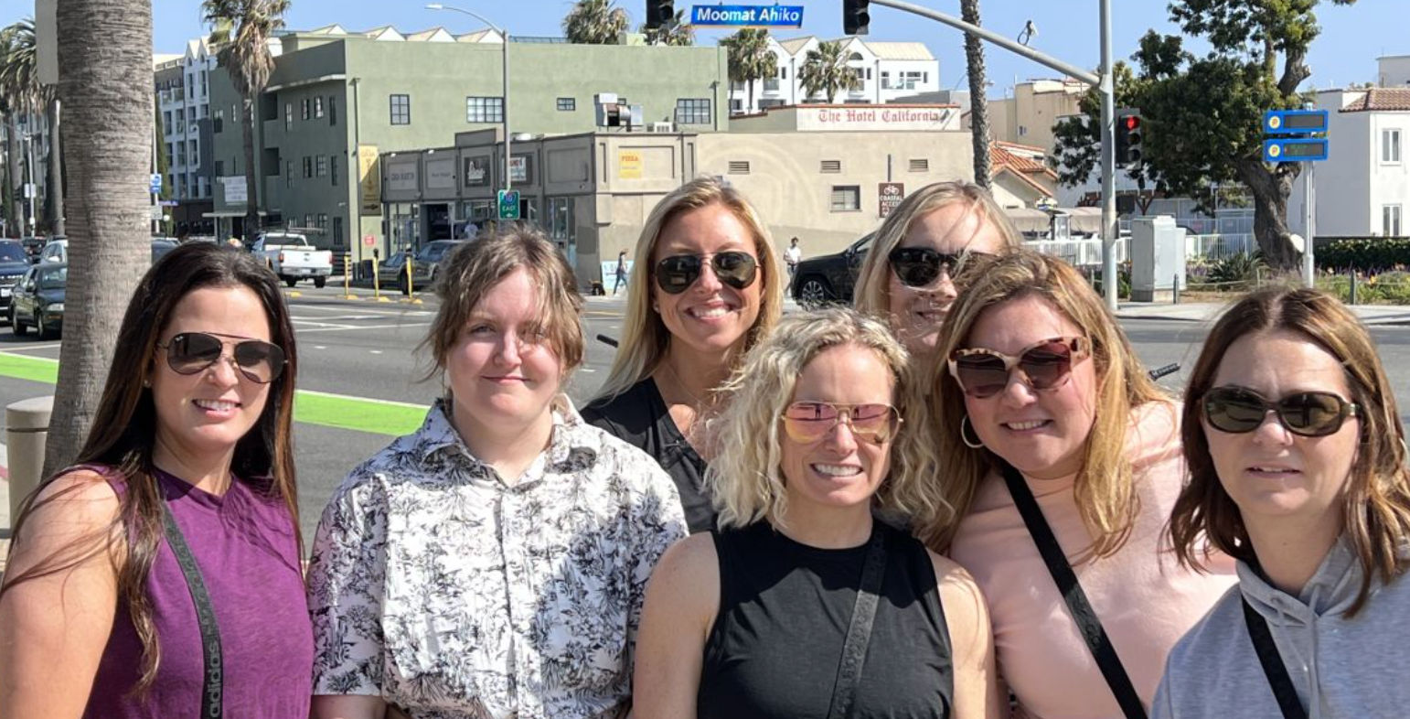 Seven smiling women wearing sunglasses pose on a sunny Southern California beachside boulevard with palm trees, cars and a green bike lane in the background.