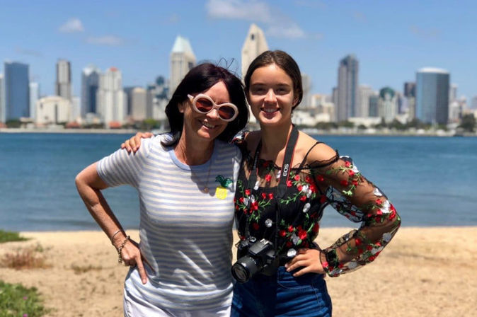 Two smiling women posing on a sunny waterfront beach with a DSLR camera, one in a floral sheer top and the other in a striped shirt and pink sunglasses, downtown skyline across the bay.