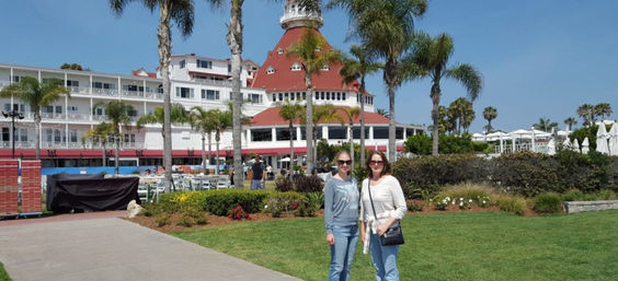 Two friends posing on a sunny lawn in front of a red-roofed Victorian seaside hotel with palm trees under a clear blue California sky.