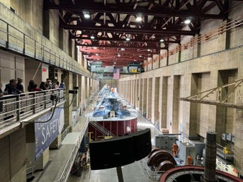 Visitors on an upper catwalk overlooking large hydroelectric turbine generators in a long concrete power plant hall with red steel roof trusses and an American flag