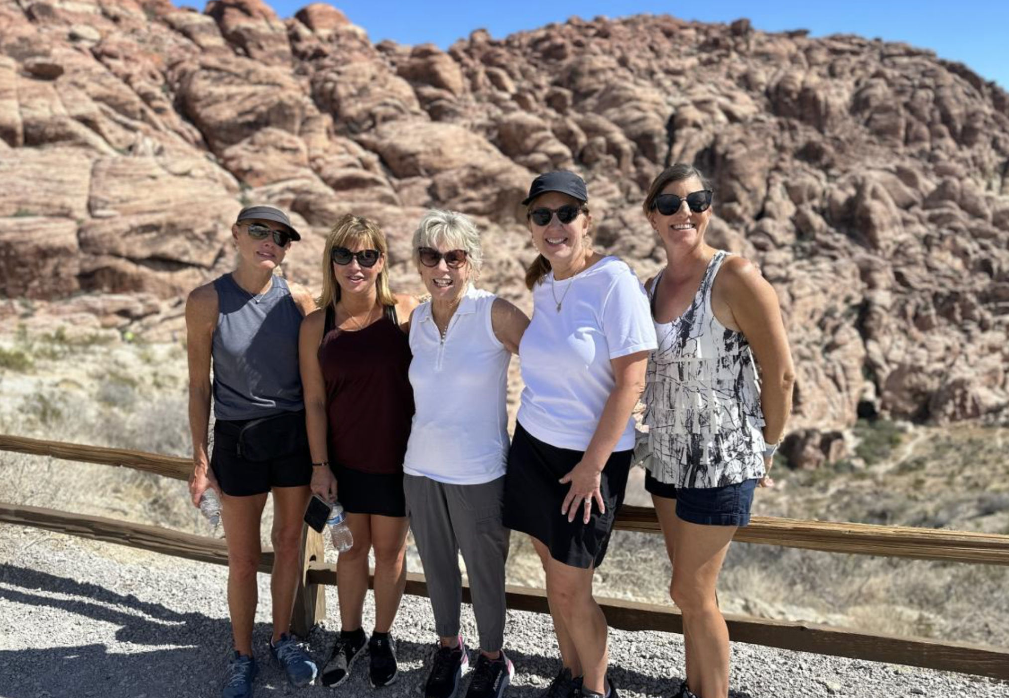 Five women hikers smiling at a scenic desert overlook with red sandstone cliffs and a clear blue sky