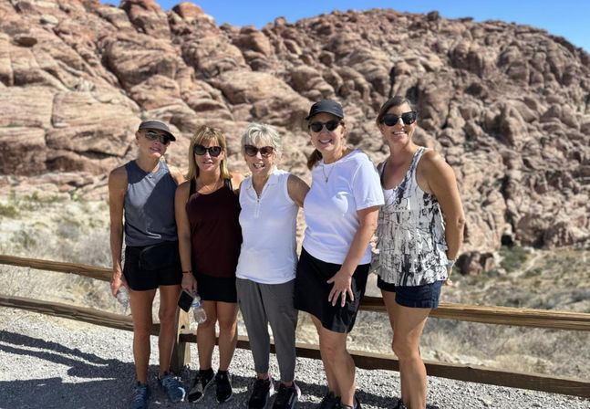Five women hikers smiling at a scenic desert overlook with red sandstone cliffs and a clear blue sky
