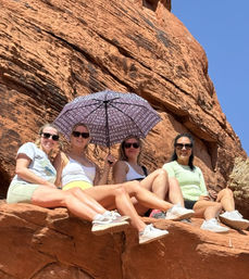 Four women in sunglasses sitting on red sandstone cliffs under a clear blue sky, one holding a patterned umbrella for shade on a sunny desert hike.