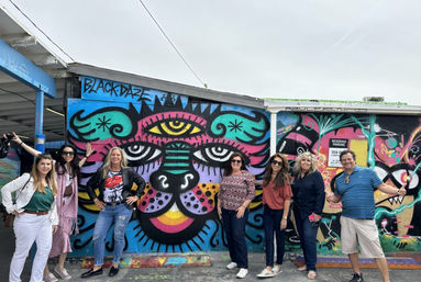 Seven adults posing in front of a vibrant street-art mural of a stylized multicolored cat face painted on an outdoor urban wall by a parking area
