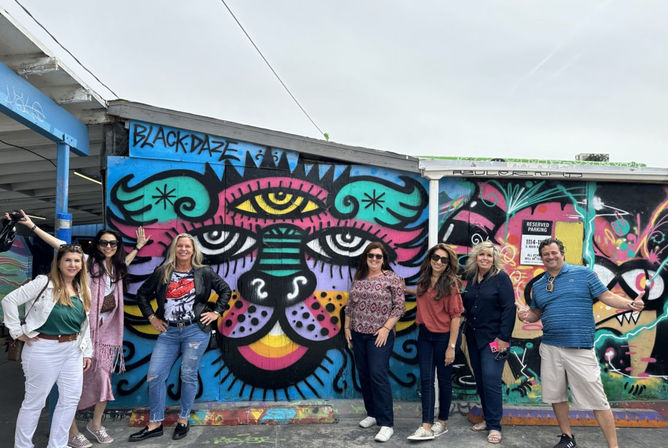 Seven adults posing in front of a vibrant street-art mural of a stylized multicolored cat face painted on an outdoor urban wall by a parking area