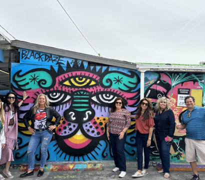 Seven adults posing in front of a vibrant street-art mural of a stylized multicolored cat face painted on an outdoor urban wall by a parking area