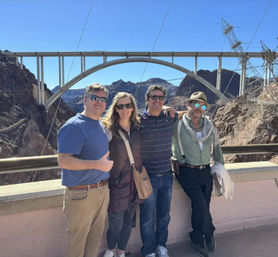 Four adults posing on a Hoover Dam overlook with the arched Mike O'Callaghan-Pat Tillman Memorial Bridge and rocky desert canyon in the background under a clear blue sky.