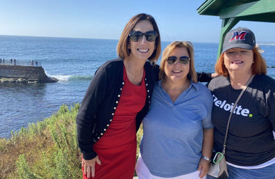 Three smiling women wearing sunglasses posing at a sunny ocean bluff viewpoint with blue sea, rocky jetty and coastal vegetation in the background.