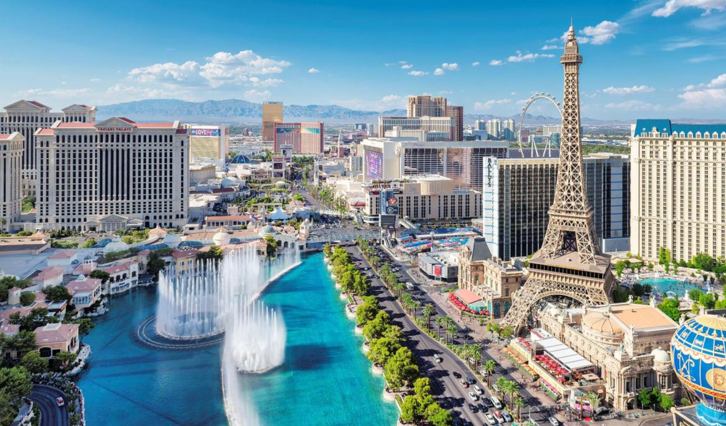 Aerial view of a vibrant sunny city strip with a turquoise fountain lake shooting choreographed jets, a Paris-style iron tower, rows of high-rise hotels, a palm-lined boulevard and a distant Ferris wheel with mountains beyond.