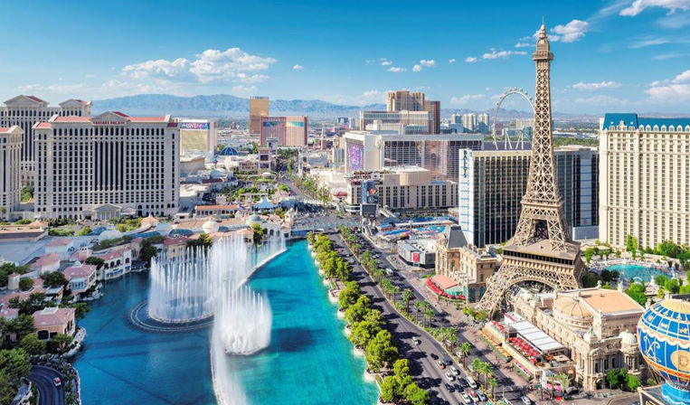 Aerial view of a vibrant sunny city strip with a turquoise fountain lake shooting choreographed jets, a Paris-style iron tower, rows of high-rise hotels, a palm-lined boulevard and a distant Ferris wheel with mountains beyond.