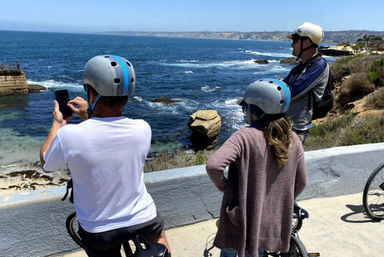 Three cyclists in helmets pause on an oceanfront bike path, one taking a photo of the rocky shoreline and deep-blue sea under a clear sky.