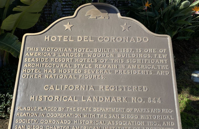 Bronze California historical landmark plaque describing a Victorian seaside hotel built in 1887, displayed outdoors among palm and bird-of-paradise plants.