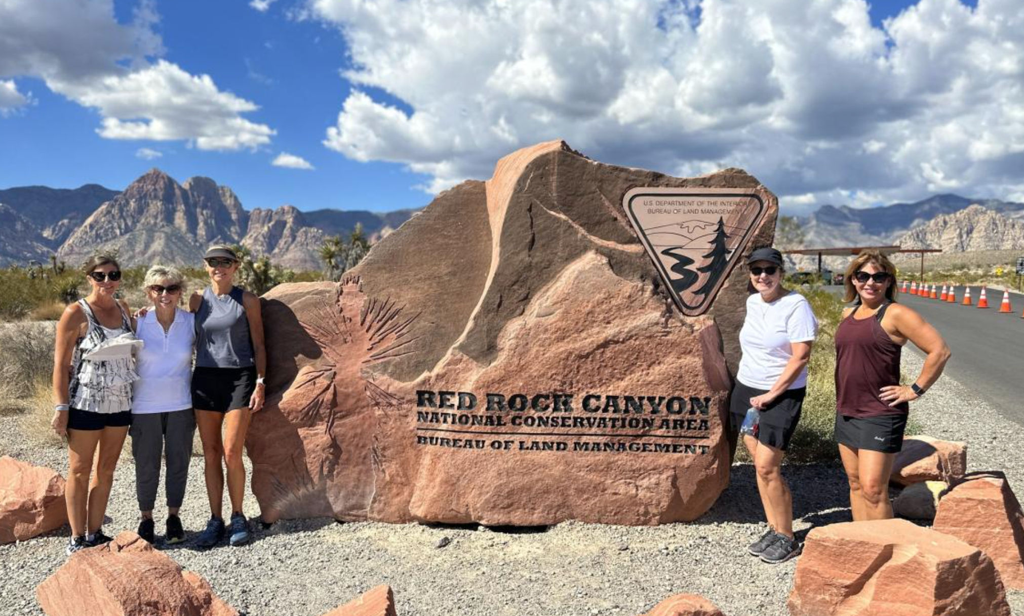 Five women hikers posed beside a large red sandstone sign reading Red Rock Canyon National Conservation Area, with desert scrub, rugged sandstone peaks and a bright blue sky.