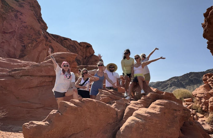 Group of hikers posing and cheering on red sandstone formations in a sunlit desert canyon under a clear blue sky