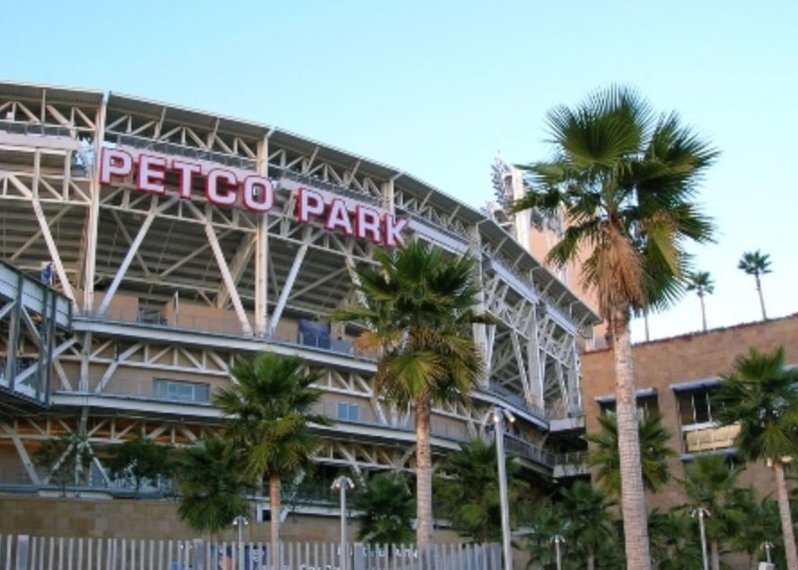 Downtown San Diego baseball stadium exterior with exposed steel trusses and red rooftop signage, palm trees in the foreground and a clear blue sky for a sunny coastal vibe.