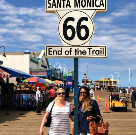 Santa Monica Pier, California — Route 66 "End of the Trail" sign on a sunny boardwalk with colorful market umbrellas, shops and visitors.