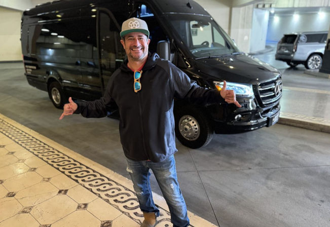 Smiling man in a cap and sunglasses hanging from his jacket, flashing shaka signs while posing in front of a black passenger van at a hotel covered valet/drop-off area with patterned tile flooring.