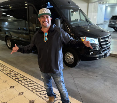 Smiling man in a cap and sunglasses hanging from his jacket, flashing shaka signs while posing in front of a black passenger van at a hotel covered valet/drop-off area with patterned tile flooring.