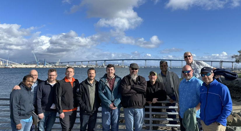 Cheerful group of men posing on a San Diego waterfront railing with the Coronado Bridge, harbor cranes and a bright blue sky with fluffy clouds.