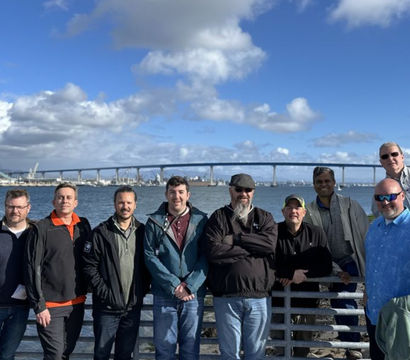 Cheerful group of men posing on a San Diego waterfront railing with the Coronado Bridge, harbor cranes and a bright blue sky with fluffy clouds.