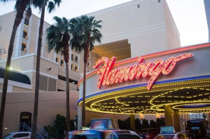 Dusk scene of a retro neon pink marquee and glowing canopy at a Las Vegas hotel entrance, tall palm trees and taxis in the foreground.