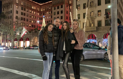 Group of four bundled in winter coats smiling on a downtown crosswalk at night in front of an ornate, festively lit building with holiday trees, decorative lights, red awnings and passing cars.