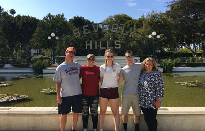 Five adults smiling and posing side-by-side in front of the Beverly Hills arch over a lily-pad pond, with trees and blue sky in Beverly Hills, Los Angeles.