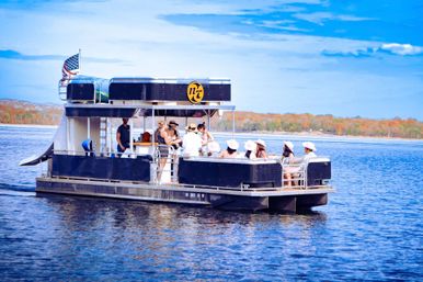 Passengers enjoying a two-level pontoon party boat flying an American flag, cruising on a blue lake past a tree-lined shoreline with autumn foliage under a bright sky