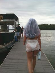 Bride-to-be in a white veil and lace outfit walking on a lakeside dock toward a pontoon boat during a bachelorette party, holding a water bottle under an overcast sky.