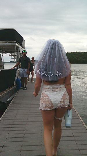 Bride-to-be wearing a veil and lace outfit walking barefoot on a wooden lake dock toward a pontoon boat and waterfront under an overcast sky