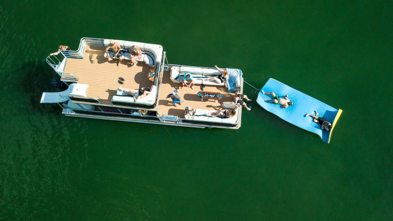 Aerial view of a pontoon boat on a green lake with people lounging on the deck and sunbathing on a blue inflatable mat tied to the side — summer boating and lake relaxation.
