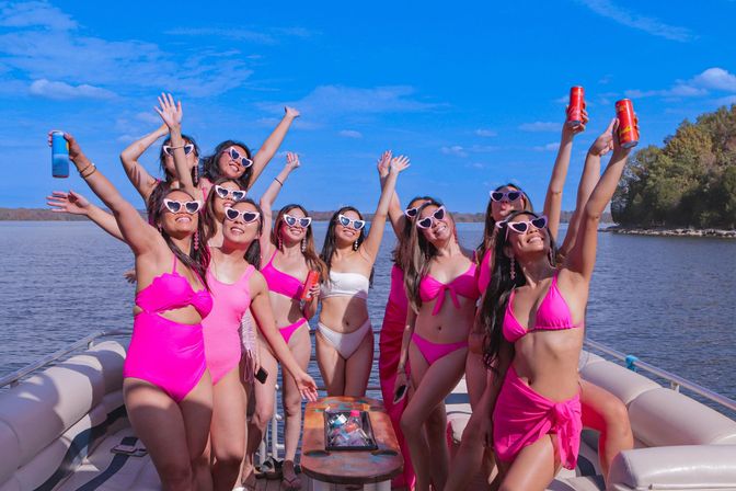 Group of friends in pink swimsuits and heart-shaped sunglasses cheering with drinks on a pontoon boat under a sunny blue sky on a lake.