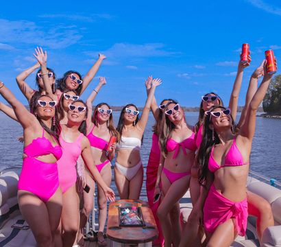 Group of friends in pink swimsuits and heart-shaped sunglasses cheering with drinks on a pontoon boat under a sunny blue sky on a lake.