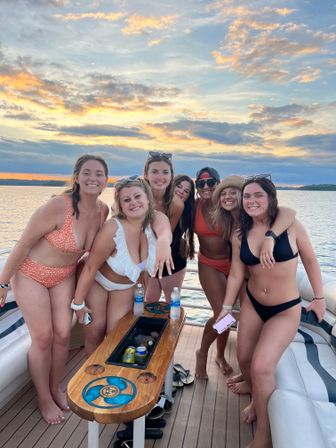 Group of seven friends in swimsuits on a boat deck at a lake sunset, smiling around a wooden drink table — summer boat outing with golden sky and calm water.