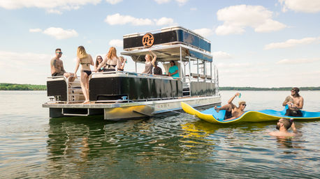 Friends enjoying a sunny summer lake party on a two-level pontoon boat, some onboard and others on yellow paddleboards in calm water with a tree-lined horizon.