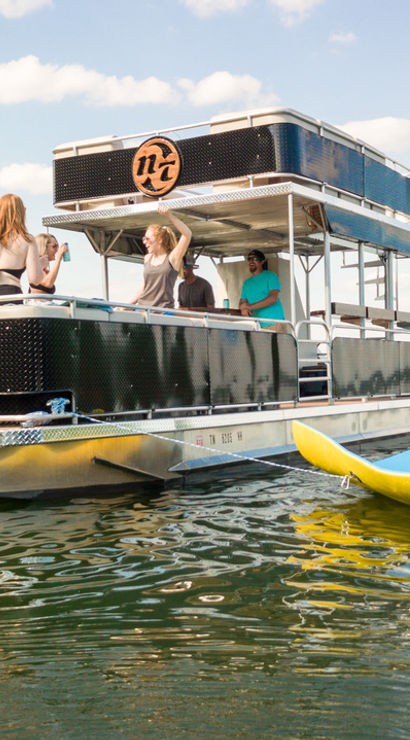 Friends enjoying a sunny summer lake party on a two-level pontoon boat, some onboard and others on yellow paddleboards in calm water with a tree-lined horizon.