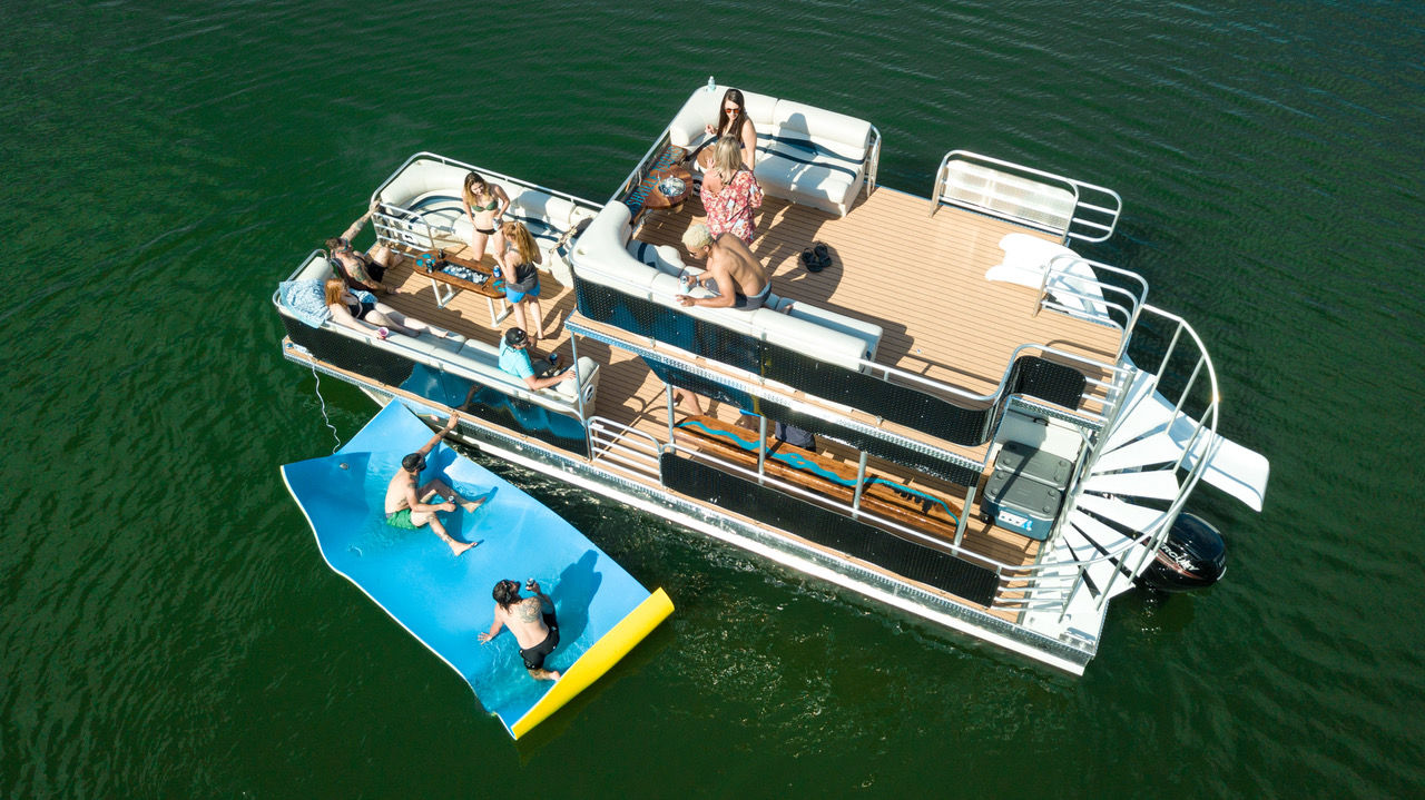Aerial drone view of a double-deck pontoon boat on a green lake, people socializing on the sunlit deck and swimmers on a bright blue inflatable slide beside the boat — lively summer boating scene.