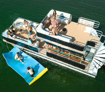 Aerial drone view of a double-deck pontoon boat on a green lake, people socializing on the sunlit deck and swimmers on a bright blue inflatable slide beside the boat — lively summer boating scene.