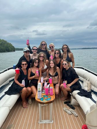 Group of smiling women on a pontoon boat on a lake, wearing swimsuits and holding pink drink cans around a small wooden table under an overcast sky.