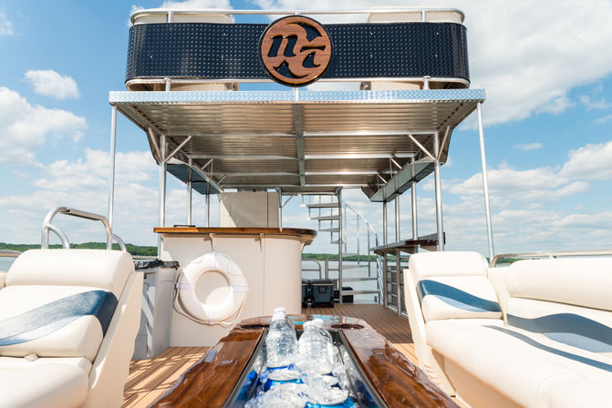 Double-decker pontoon boat deck on a sunny lake with white-and-blue cushioned seating, center ice-filled cooler of water bottles, bar area, life ring and spiral staircase to the upper deck.