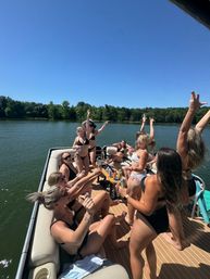 Summer lake party on a pontoon boat with women in swimsuits raising drinks and hands, tree-lined shoreline and clear blue sky