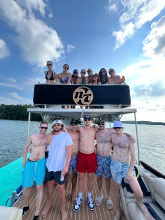 Group of friends on a pontoon boat enjoying a sunny lake day, people in swimsuits posing on upper and lower decks with blue sky and tree-lined shore