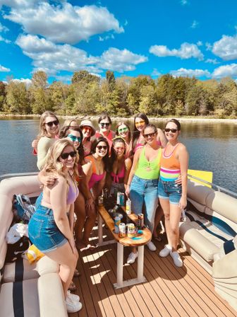 Group of friends on a pontoon boat at a sunny lake, smiling in colorful swimsuits and sunglasses around a table with drinks, tree-lined shoreline and blue sky with fluffy clouds.
